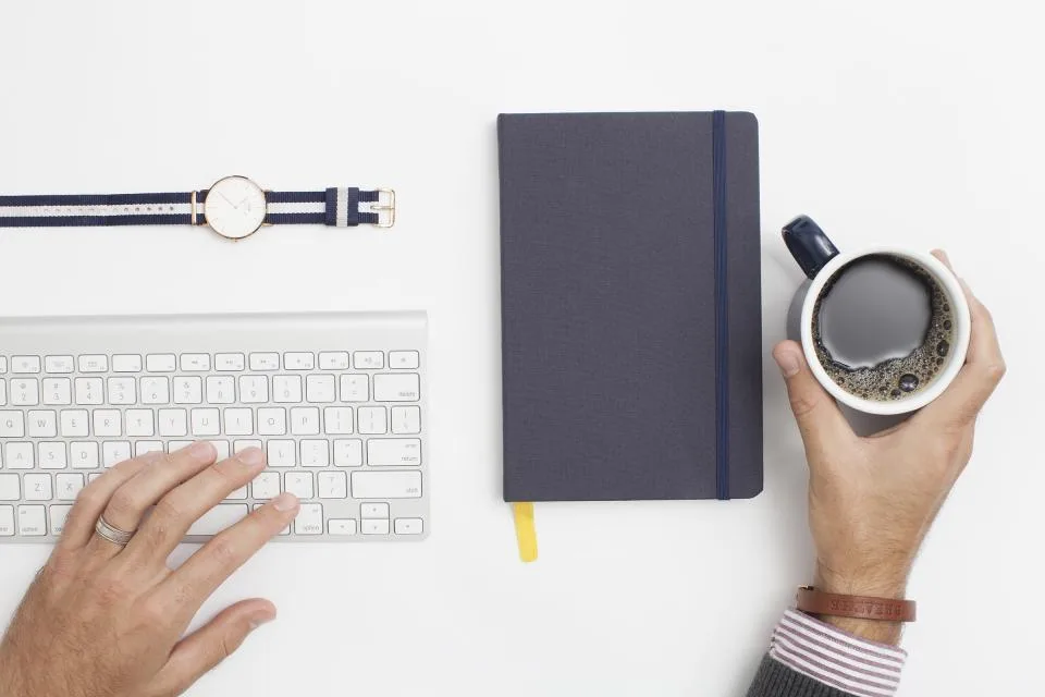 Hands at a white keyboard beside a dark notebook and a cup of coffee on a clean desk