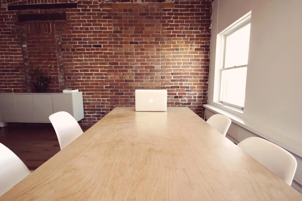 Bright meeting room with a long wooden table, white chairs and a laptop against a brick wall