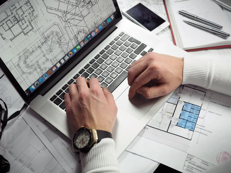 Hands typing on a laptop surrounded by printed plans and documents on a white desk