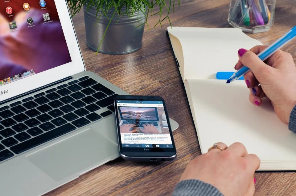 Laptop, smartphone and an open notebook on a wooden desk with a hand writing notes