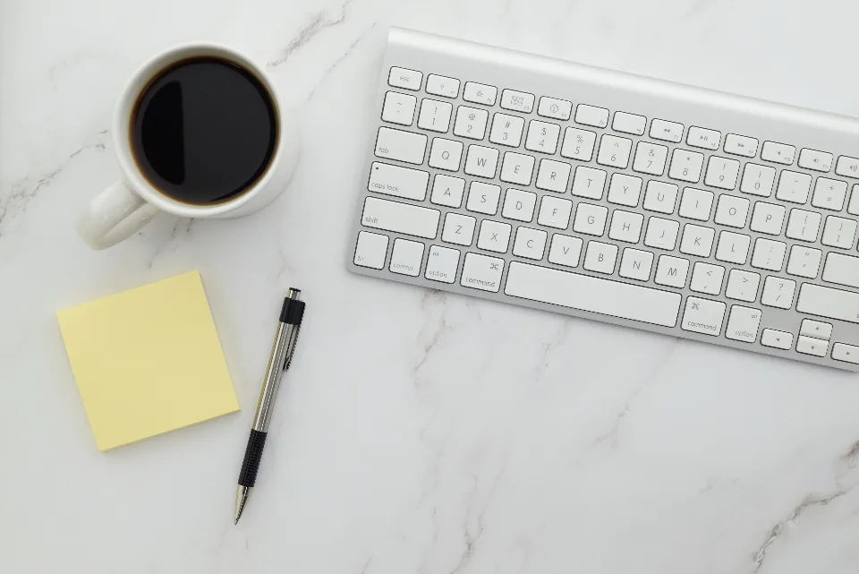 White keyboard, cup of black coffee, yellow sticky note and pen on a marble desk