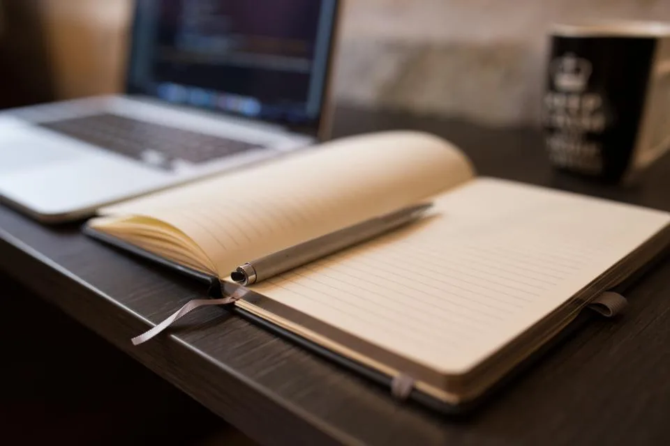 Open lined notebook with a pen beside a laptop on a desk