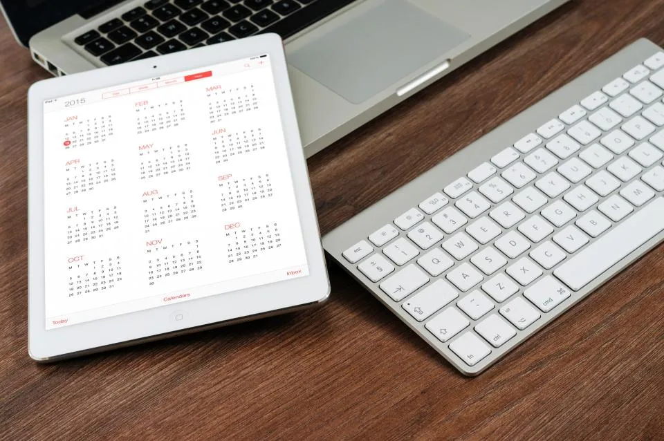 Tablet showing a calendar next to a white keyboard on a wooden desk