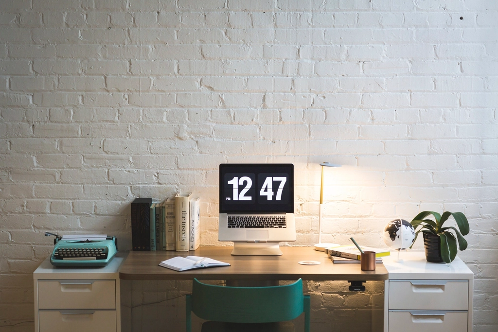 Tidy home office desk against a white brick wall with a computer, typewriter, books and a plant