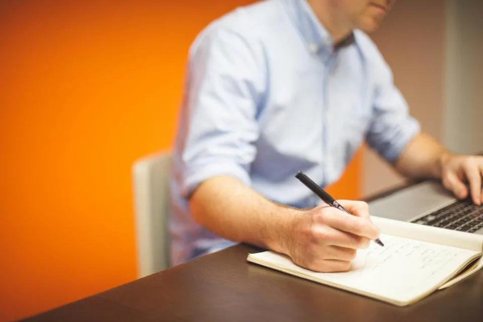 Person in a light blue shirt writing notes in a notebook beside a laptop